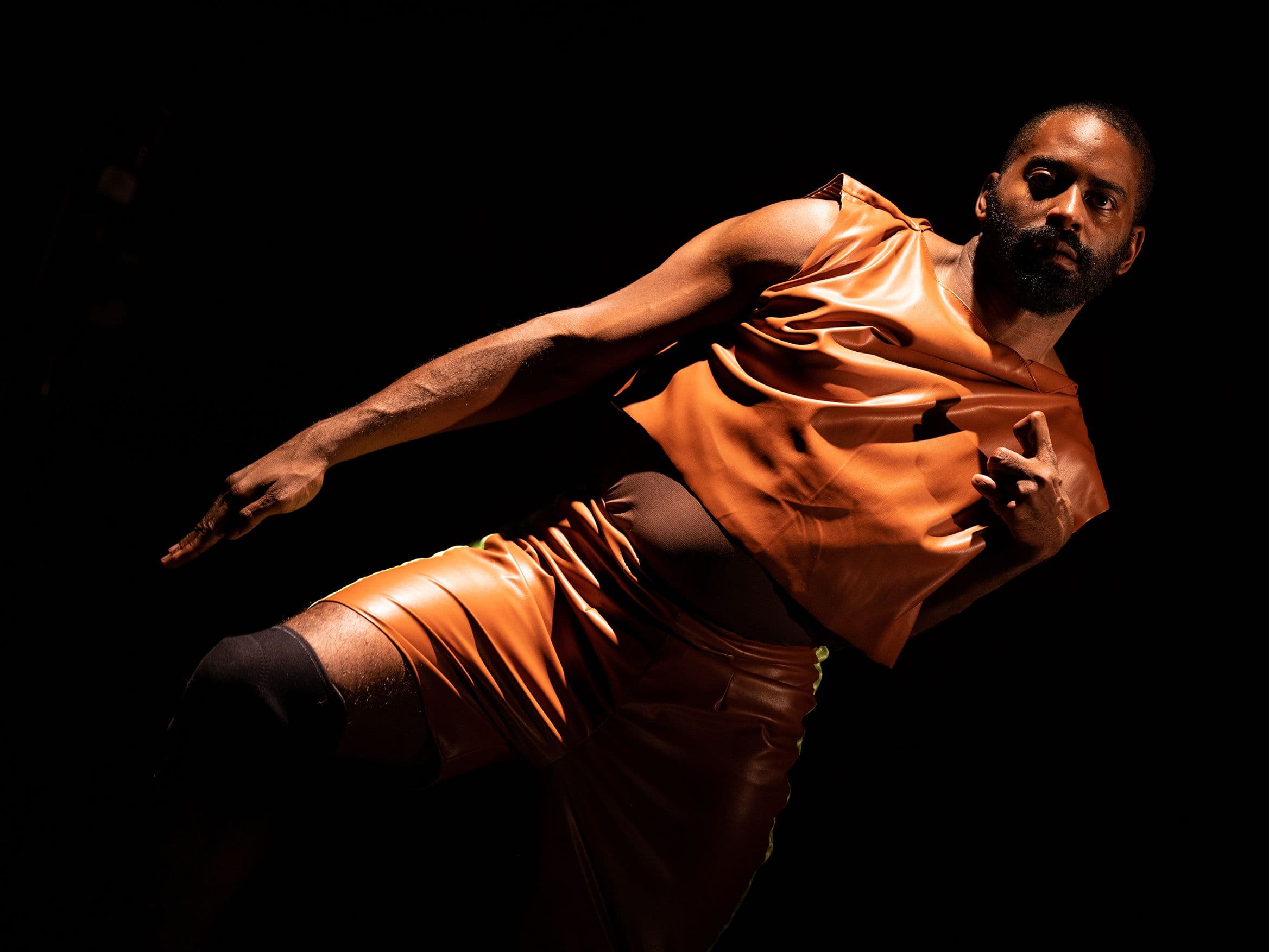 Jerron, a dark-skinned Black man leans with arm outstretched in a dark orange tunic By Maria Baranova